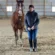 A woman wearing a dark jacket, jeans, and boots stands indoors on dirt beside a brown horse, holding its reins. The background shows wooden beams and metal siding.