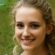 A young woman with wavy blonde hair smiles at the camera. She has blue eyes and is wearing a light top. The blurred green background suggests an outdoor setting, perfect for a fresh 2024 Sale Page or Training Scale promotion.