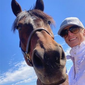 Member Becky, wearing sunglasses and a cap, smiles while standing next to a dark brown horse under a bright blue sky. Captured from a low angle, the horse’s powerful strides make it appear even larger against the scattered clouds.