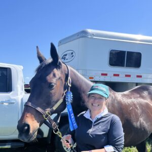 A smiling woman in a green cap holds a dark brown horse with a blue ribbon on its bridle, standing in front of a white truck and horse trailer on a sunny day.