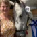 A woman in a yellow floral top smiles beside a gray horse wearing a bridle and number tag 11, with its head leaning affectionately on her. Blue and white ribbons hang in the background.