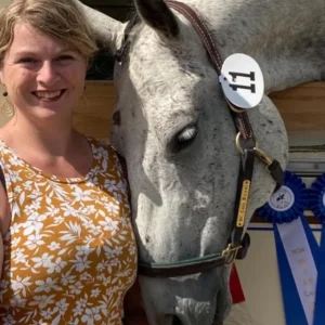 A woman in a yellow floral top smiles next to a gray horse wearing a bridle and number tag. The horse rests its head gently on her shoulder. Blue ribbons are visible in the background.