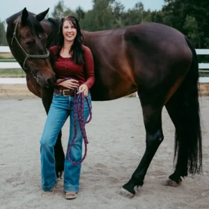 A woman wearing a red long-sleeve shirt and blue jeans stands smiling next to a dark brown horse in an outdoor fenced area, holding a purple and pink lead rope. Trees are visible in the background.