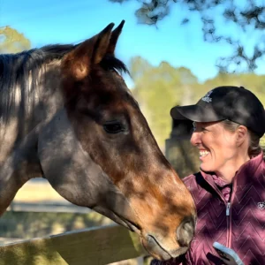 A woman in a maroon jacket and black cap smiles at a brown horse, standing close together by a wooden fence on a sunny day, with trees and blue sky in the background.