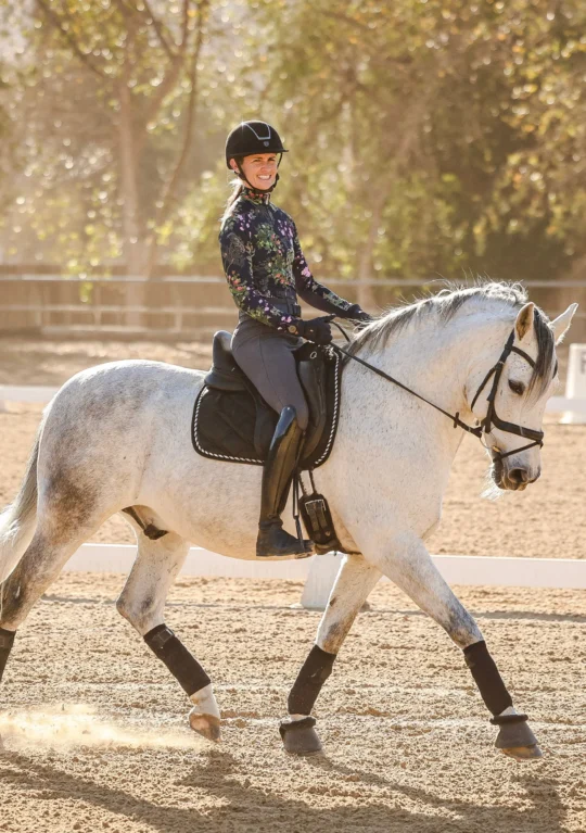 A person wearing a helmet and floral-patterned shirt smiles while riding a white horse in an outdoor sandy arena, surrounded by trees in the background.