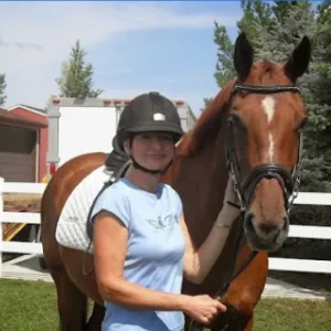 A woman wearing a helmet and blue shirt stands next to a brown horse with a white saddle and bridle, holding its reins in a grassy area with a white fence, trees, and a trailer in the background.