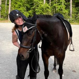 A person wearing a helmet stands close to a dark brown horse, smiling, in an outdoor sandy area with trees in the background. The horse is saddled and gently nuzzling the person.