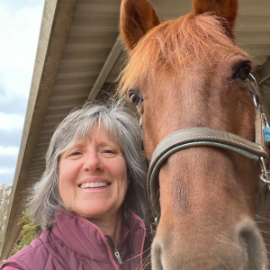 A smiling woman with gray hair takes a close-up selfie with a brown horse wearing a bridle, both under a covered outdoor structure, showcasing the groundwork of their bond.